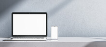 Close up of empty white laptop computer and smartphone on gray desk. Concrete wall background. Device presentation and online education concept. Mock up, 3D Rendering.