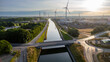 This aerial image captures a modern landscape at dawn, where renewable energy meets waterway transportation. Wind turbines line the canal, symbolizing a commitment to sustainable energy, while the