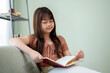 © bongkarn - A cute and happy young Asian girl in casual clothes is reading a book on a sofa in the living room.