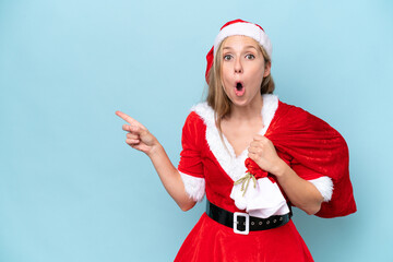 Wall Mural - Young blonde woman dressed as Mama Claus holding a sack isolated on blue background surprised and pointing side