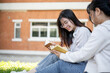 © bongkarn - A female student is reading a book while relaxing under the tree in a campus park with her friend.