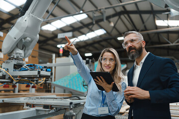 Wall Mural - Female engineer and male project manager standing in modern industrial factory by precision robotic arm. Robot assembling wooden furniture in big furniture manufacturing facility with industrial
