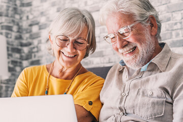 Pretty elderly 70s grey-haired couple resting on couch in living room hold on lap laptop watching movie smiling enjoy free time, older generation and modern wireless technology advanced users concept.