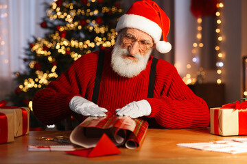 Poster - Santa Claus wrapping gift at his workplace in room decorated for Christmas