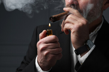 Bearded man lighting cigar on dark grey background, closeup