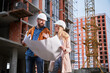 Couple in safety helmets studying architectural drawings outside apartment building under construction. Man and woman discussing building plan outdoors at construction site.