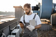 © sofiko14 - Focused bearded master dressed in overalls standing and holding tablet while using digital smartphone on fresh air. Caucasian skilled man using gadgets and maintaining equipment on roof of factory.
