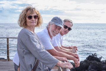 Sticker - Group of relaxed mature and senior friends standing face the sea at sunset light, three people looking at camera enjoying freedom and vacation or retirement