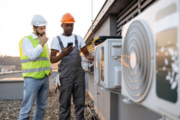 Team of two multiracial technicians checking voltage with modern multimeter and discussing repairing of cooling system. Competent male inspectors with toolbox looking at air conditioner outdoors.