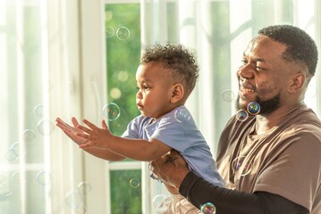 Wall Mural - Happy african black parents dad father throw baby son on piggyback and neck riding teasing fun near window. Black baby son and daddy enjoy teasing kiss cheek and neck riding at window light