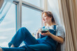 © okrasiuk - Free Time To Relax. Young woman looks out the window overlooking the city, sits on the windowsill at cozy home holds cup of hot tea drink. Happy calm female taking break for mental health wellbeing