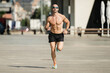 © carlesiturbe - Athletic young man running in the street wearing sunglasses. Front view of sportsman jogging outdoors shirtless.