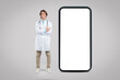© Prostock-studio - Young male doctor in white coat standing near huge smartphone with white blank screen over grey background, mockup
