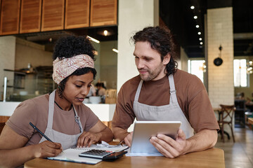 Two biracial eatery workers sitting over income calculation sitting at table after workday