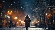 A woman is seen walking down a snowy street at night. This image can be used to depict a winter night scene or a peaceful evening stroll.