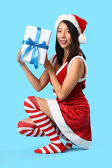 Poster - Young Asian woman in Santa costume with Christmas gift on blue background