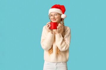 Wall Mural - Beautiful young happy woman in Santa hat with cup of tea on blue background