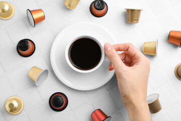 Poster - Woman holding cup of coffee at white tiled table with capsules, top view