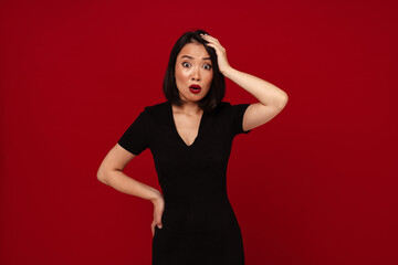 Wall Mural - Shocked asian woman grabbing her head while standing isolated over red wall