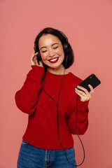 Wall Mural - Cheerful woman listening music with headphones and mobile phone while standing isolated over pink background