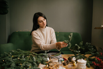 Wall Mural - Woman making Christmas wreath using natural pine branches, sitting near wooden table.