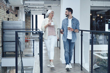 Smiling man ans woman holding coffee while walking in the office hall, talking, having coffee break. Full-length. Teamwork, startup, business concept