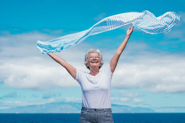 Sticker - Cheerful attractive senior woman with outstretched arms enjoying sunny day, freedom and sea vacation waving scarf in the wind. Happy retirement concept