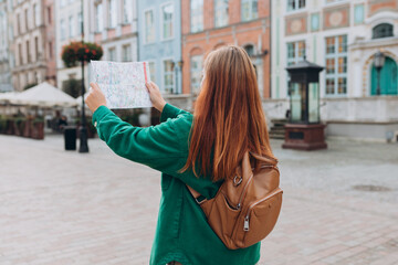 Wall Mural - Attractive young female tourist is exploring city. Redhead 30s woman with backpack holding a paper map on city street. Traveling Europe in autumn, rear view