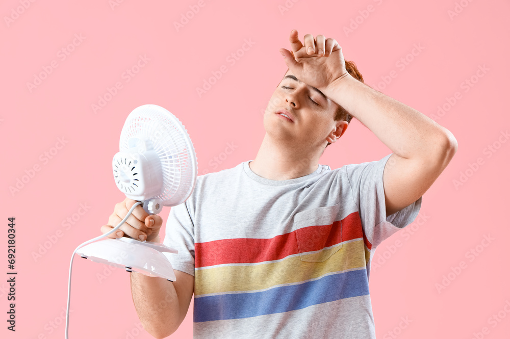 Sweaty young man with electric fan on pink background