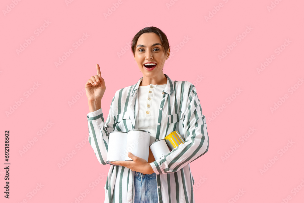 Young woman with paint cans pointing at something on pink background