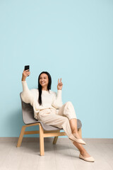 Sticker - Beautiful young woman sitting on comfortable chair near blue wall and taking selfie