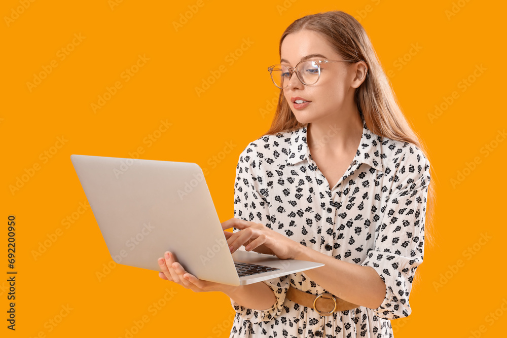 Young woman in eyeglasses using laptop on yellow background