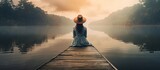 Fototapeta  - Back view of fashioned young woman sitting on wooden dock looking at view on a misty morning Female hipster with brown hat relaxes on the edge of jetty admiring foggy lake Wonderful nature geta