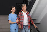 Fototapeta  - Young healthcare worker assisting senior woman on stairs indoors