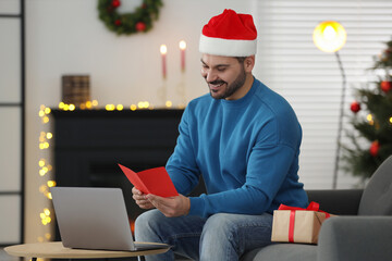 Poster - Celebrating Christmas online with exchanged by mail presents. Smiling man in Santa hat reading greeting card during video call on laptop at home