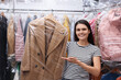 Dry-cleaning service. Happy woman holding hanger with coat in plastic bag near racks with clothes indoors