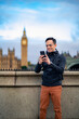 Positive young Latin male tourist standing on embankment and using smartphone in front of historic building located in London near Big Ben against clock tower during holiday