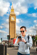 Young cheerful Latin man tourist wearing sunglasses and casual outfit holding smartphone while standing against Big Ben and clock tower barrier and smiling in London