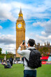Unrecognizable young cheerful Latin man tourist wearing sunglasses and casual outfit holding smartphone while standing against Big Ben and clock tower barrier and smiling in London