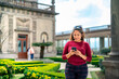 Happy young Latin woman tourist in casual clothes and sunglasses browsing smartphone while standing against blurred historic Chapultepec Castle in Mexico City
