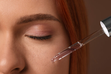 Wall Mural - Beautiful young woman applying cosmetic serum onto her face on brown background, closeup