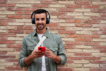 Wall Mural - Portrait of a handsome charming man isolated in a brick wall, using smartphone to listen music wearing headphones, looking at camera smiling. Copy space.