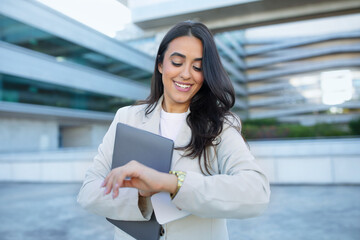 Poster - Latin entrepreneur woman smiling checking watch standing with laptop outdoor
