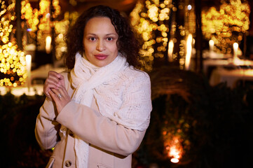 Wall Mural - Attractive young woman looking confidently at camera standing against Christmas lights backdrop at funfair at night time