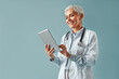 Beautiful mature female doctor in white coat holding digital tablet and looking at it, smiling while standing against blue background. Healthcare and digital technologies.