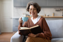 Happy elderly senior woman drinking hot tea and reading favorite paper book, enjoying cozy weekend, relaxing alone on sofa, smiling at camera