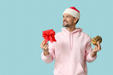 Canvas Print - Handsome man in Santa hat with gift box and tasty cookies on blue background