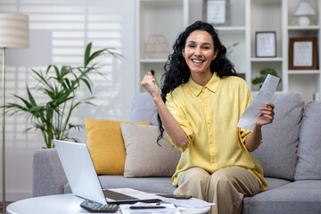 Wall Mural - Joyful hispanic businesswoman holding work documents in hands sitting on sofa, happy with good news, success at work, looking at camera.