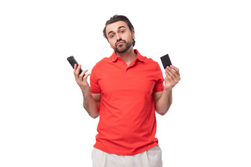 Wall Mural - young brutal man with black hair and a beard dressed in a red t-shirt with a mockup for printing holds a plastic card and a smartphone on a white background with copy space