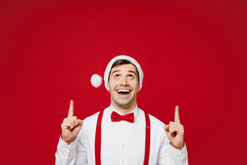 Wall Mural - Merry young smiling cool man wear white shirt Santa hat posing point index finger overhead on area mock up isolated on plain red background studio Happy New Year Christmas celebration holiday concept